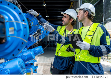 Two technicians in high-visibility vests examine blue industrial machinery with focused expressions Two technicians in high-visibility vests examine blue industrial machinery with focused expressions 141845172