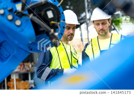Two technicians in safety gear inspect blue robotic arm's internal components in industrial workshop with focused expressions Two technicians in safety gear inspect blue robotic arm's internal components in industrial workshop with focused expressions 141845173