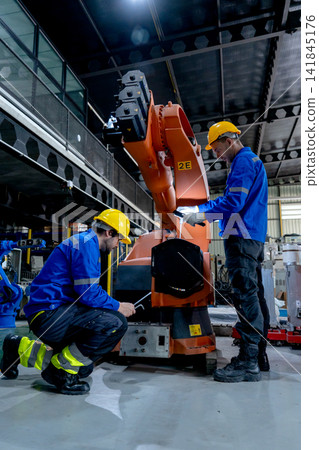 Two technicians in blue uniforms and yellow hard hats inspect orange robotic arm's joint with focused expressions in industrial workshop Two technicians in blue uniforms and yellow hard hats inspect orange robotic arm's joint with focused expressions in industrial workshop 141845176