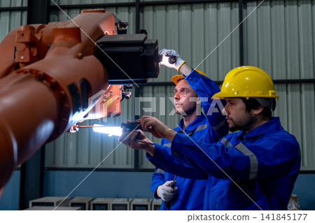 Two technicians in blue uniforms and yellow hard hats inspect orange robotic arm's joint with focused expressions in industrial workshop Two technicians in blue uniforms and yellow hard hats inspect orange robotic arm's joint with focused expressions in industrial workshop 141845177