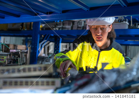 Female technician in bright yellow safety jacket examines mechanical part under blue industrial shelving Female technician in bright yellow safety jacket examines mechanical part under blue industrial shelving 141845181