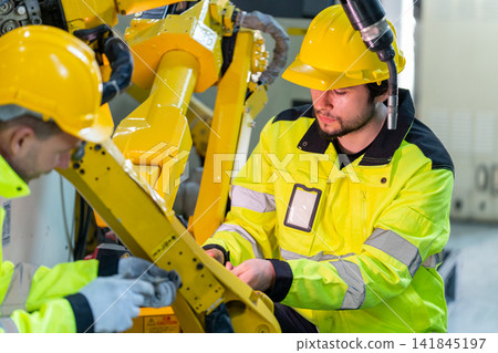 Two technicians in yellow hard hats and safety jackets adjust large robotic arm joint in industrial workshop with visible cables and tools Two technicians in yellow hard hats and safety jackets adjust large robotic arm joint in industrial workshop with visible cables and tools 141845197