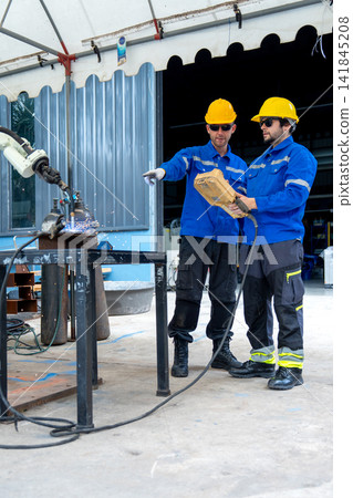 Two engineers in blue uniforms and yellow hard hats operate robotic welding arm in industrial workshop 141845208