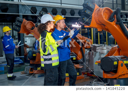 Two technicians in safety gear inspect orange robotic arm's internal components with focused expressions in modern factory 141845220