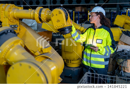 Female technician in high-visibility jacket checks yellow robotic arm in industrial warehouse with focused expression Female technician in high-visibility jacket checks yellow robotic arm in industrial warehouse with focused expression 141845221