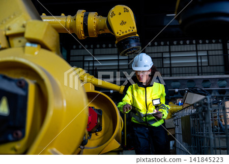 Female technician in high-vis jacket inspects yellow robotic arm's joint with focused expression in industrial workshop Female technician in high-vis jacket inspects yellow robotic arm's joint with focused expression in industrial workshop 141845223