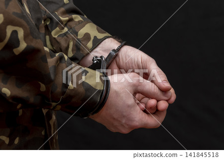 A military man in camouflage is handcuffed on a dark background. Concept: war criminal, prisoner of war, tribunal for deserters, traitor to the motherland. A military man in camouflage is handcuffed on a dark background. Concept: war criminal, prisoner of war, tribunal for deserters, traitor to the motherland. 141845518
