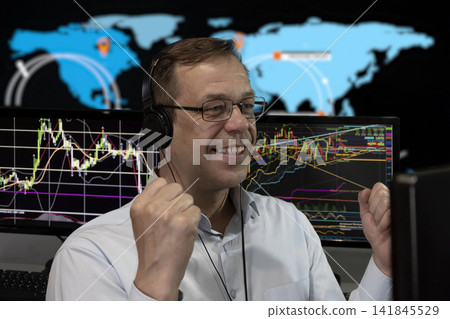 An elderly male broker rejoices in the growth of the exchange rate against the background of the stock tableau and the world map. Concept: currency exchange rate growth, online trading, increased An elderly male broker rejoices in the growth of the exchange rate against the background of the stock tableau and the world map. Concept: currency exchange rate growth, online trading, increased 141845529