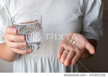 Girl in a dress take the two pills and glass with water in the hand. Healthcare from pharmacy. Kid sit at home because of illness. 141847100