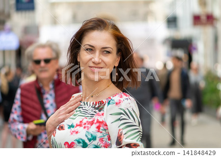 Smiling brunette woman in floral blouse glancing sideways on busy city street Smiling brunette woman in floral blouse glancing sideways on busy city street 141848429
