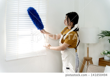 Asian adult woman cleaning dusty window blinds with blue duster at home for domestic hygiene and tidy living space 141850494