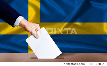 Hand placing a ballot in ballot box with the Swedish flag in the background. Sweden Parliamentary vote 2026 Hand placing a ballot in ballot box with the Swedish flag in the background. Sweden Parliamentary vote 2026 141851034