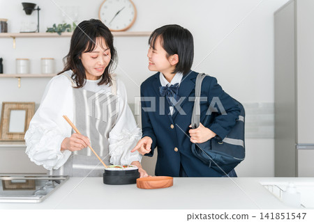早上,一位母親在廚房為女兒準備午餐,女孩穿著制服(正在做午餐)。 早上,一位母親在廚房為女兒準備午餐,女孩穿著制服(正在做午餐)。 141851547