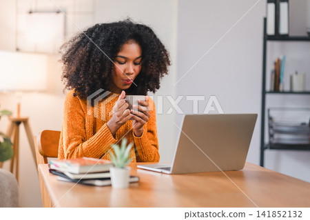 Focused African American woman holding a coffee mug while working on a laptop at home, sitting at a desk with books Focused African American woman holding a coffee mug while working on a laptop at home, sitting at a desk with books 141852132