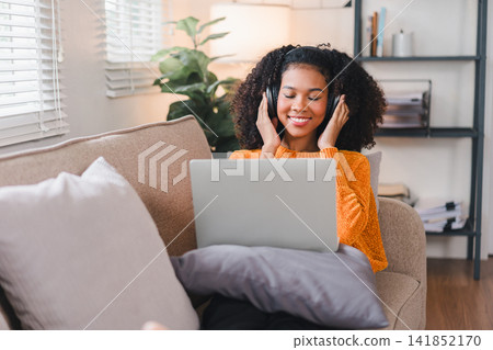Smiling African American woman relaxing on a cozy couch, wearing headphones, using a laptop, enjoying a comfortable home Smiling African American woman relaxing on a cozy couch, wearing headphones, using a laptop, enjoying a comfortable home 141852170