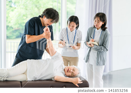 A senior woman receiving massage and stretching during rehabilitation, a male physical therapist, and a female staff member receiving training. A senior woman receiving massage and stretching during rehabilitation, a male physical therapist, and a female staff member receiving training. 141853256
