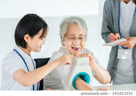 Senior woman measuring blood pressure using a sphygmomanometer and a nurse taking notes, female caregiver Senior woman measuring blood pressure using a sphygmomanometer and a nurse taking notes, female caregiver 141853331