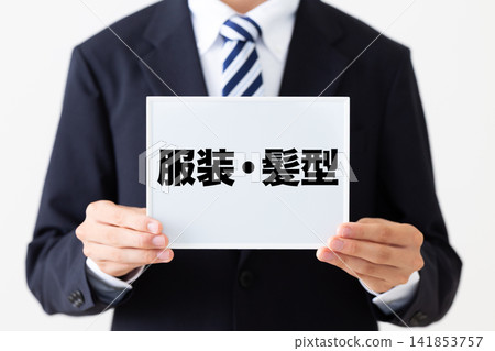 身穿西裝的年輕商人,手持一塊寫有服裝規範和髮型指南的白板。 身穿西裝的年輕商人,手持一塊寫有服裝規範和髮型指南的白板。 141853757