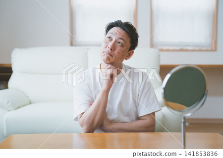 A 40-year-old man leans on his hand in front of a tabletop mirror, contemplating his blemishes during a skin check. A 40-year-old man leans on his hand in front of a tabletop mirror, contemplating his blemishes during a skin check. 141853836