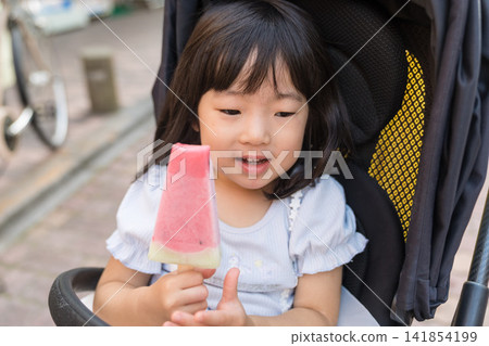 A Japanese toddler girl sitting in a stroller and munching on watermelon. A Japanese toddler girl sitting in a stroller and munching on watermelon. 141854199