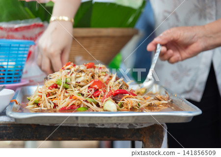 Fresh spicy Thai papaya salad tomato chili long beans savory dressing served metal tray while vendors prepare traditional street food Fresh spicy Thai papaya salad tomato chili long beans savory dressing served metal tray while vendors prepare traditional street food 141856509
