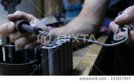 a lithium-ion battery being soldered. The technician holds a soldering iron in his left hand, pointing its tip toward the battery terminal. With his right hand, he applies thick tin solder to the join a lithium-ion battery being soldered. The technician holds a soldering iron in his left hand, pointing its tip toward the battery terminal. With his right hand, he applies thick tin solder to the join 141857847