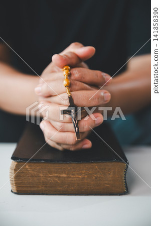 Close-up of clasped hands holding rosary beads and a crucifix above an old Holy Bible, representing Catholic prayer, Christian faith, devotion, hope, forgiveness, and salvation. Close-up of clasped hands holding rosary beads and a crucifix above an old Holy Bible, representing Catholic prayer, Christian faith, devotion, hope, forgiveness, and salvation. 141858390