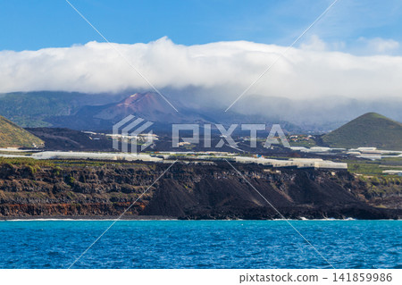 Volcanic cone of Tajogaite volcano visible on the horizon above clouds, shot from the sea near the lava delta in La Palma, Canary Islands, Spain Volcanic cone of Tajogaite volcano visible on the horizon above clouds, shot from the sea near the lava delta in La Palma, Canary Islands, Spain 141859986
