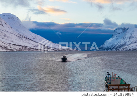 Small motorboat speeding through the cold waters of Ersfjorden fjord leaving a wake trail against snowy mountains, Ersfjordbotn, Tromso, Norway Small motorboat speeding through the cold waters of Ersfjorden fjord leaving a wake trail against snowy mountains, Ersfjordbotn, Tromso, Norway 141859994