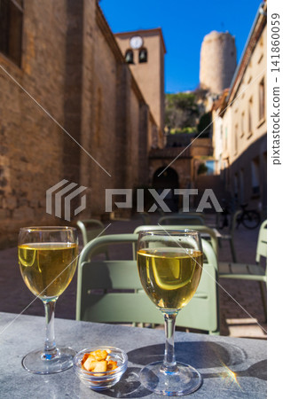 Two glasses of white wine on a table on a scenic street with the historic church and the medieval Barbarossa Tower in the background, Gruissan, Occitanie, France Two glasses of white wine on a table on a scenic street with the historic church and the medieval Barbarossa Tower in the background, Gruissan, Occitanie, France 141860059