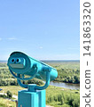 Turquoise coin-operated binocular viewer on a scenic viewpoint overlooking a river and green landscape under a blue sky. Travel and tourism concept. 141863320