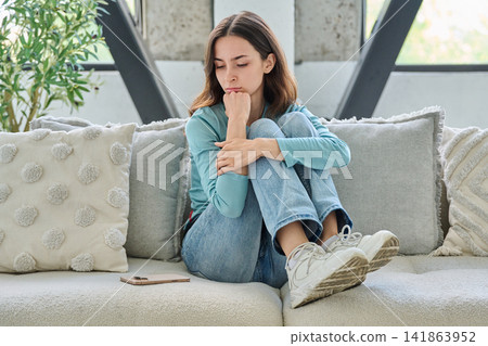 Sad young woman sitting on couch feeling anxious and lonely Sad young woman sitting on couch feeling anxious and lonely 141863952