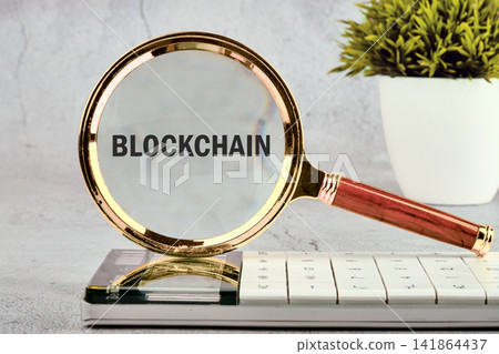 A magnifying glass highlights the word blockchain placed on a calculator. A small green plant adds to the scene A magnifying glass highlights the word blockchain placed on a calculator. A small green plant adds to the scene 141864437