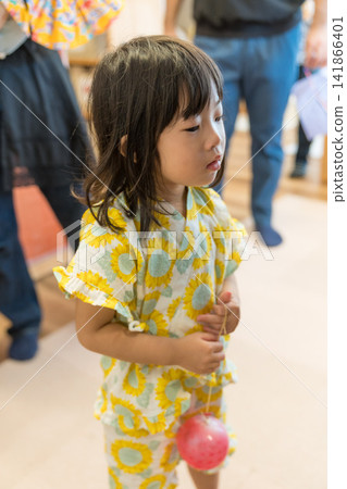 A Japanese toddler girl wearing a sunflower-patterned jinbei (traditional Japanese summer wear) and holding a water balloon. A Japanese toddler girl wearing a sunflower-patterned jinbei (traditional Japanese summer wear) and holding a water balloon. 141866401