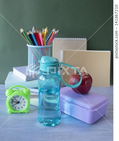 Bottle of water and lunchbox on table with school supplies. Alarm clock, textbooks, colored pencils, notebooks and healthy snack on blackboard background, back to school concept. Bottle of water and lunchbox on table with school supplies. Alarm clock, textbooks, colored pencils, notebooks and healthy snack on blackboard background, back to school concept. 141867210