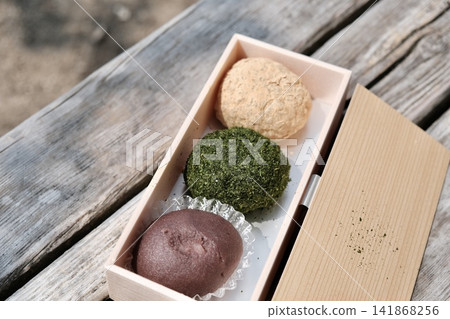 在戶外的木凳上，三個不同顏色的精緻手工萩餅（年糕）被精美地擺放在便當盒裡。 141868256