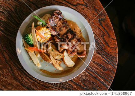 Grilled chicken with potato wedges, corn, broccoli, baby corn, carrots and brown gravy served on a white plate over a rustic wooden table, photographed from above as a restaurant meal. Grilled chicken with potato wedges, corn, broccoli, baby corn, carrots and brown gravy served on a white plate over a rustic wooden table, photographed from above as a restaurant meal. 141868967
