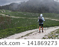 Female hiker in blue hoodie walking along scenic mountain trail with backpack and trekking poles Female hiker in blue hoodie walking along scenic mountain trail with backpack and trekking poles 141869273