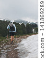 Female hiker in blue hoodie walking along scenic mountain trail with backpack and trekking poles Female hiker in blue hoodie walking along scenic mountain trail with backpack and trekking poles 141869289