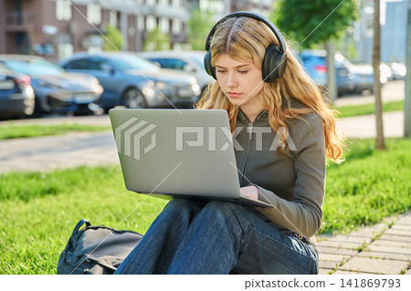 Young woman in headphones working on laptop outdoors sitting on green grass Young woman in headphones working on laptop outdoors sitting on green grass 141869793