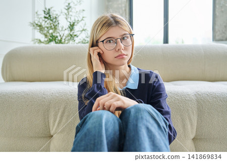 Sad teenage student girl in glasses sitting on floor hugging knees Sad teenage student girl in glasses sitting on floor hugging knees 141869834