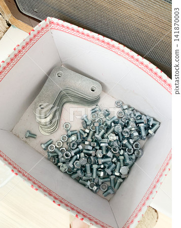 A cardboard box filled with metallic corner brackets and small steel bolts on a flat surface A cardboard box filled with metallic corner brackets and small steel bolts on a flat surface 141870003