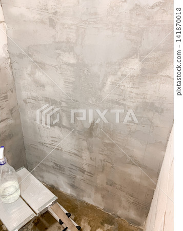 A grey cement wall with textured plaster finish stands next to a small white stepladder and a water spray bottle illustrating home renovation progress and interior decoration preparation in a room A grey cement wall with textured plaster finish stands next to a small white stepladder and a water spray bottle illustrating home renovation progress and interior decoration preparation in a room 141870018