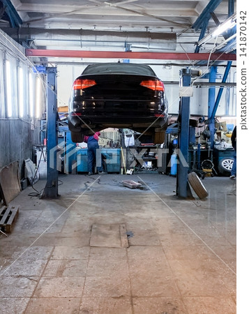A black car is lifted on a mechanical platform for repairs inside a professional auto service shop 141870142