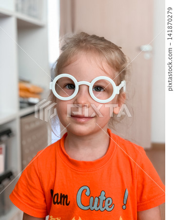 A smiling little girl with blonde hair wears oversized white circular play glasses while standing at home capturing a cute and joyful moment of imaginative dress up and innocent childhood play fun A smiling little girl with blonde hair wears oversized white circular play glasses while standing at home capturing a cute and joyful moment of imaginative dress up and innocent childhood play fun 141870279