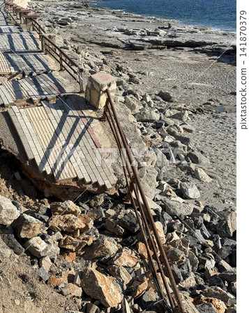 Collapsed stone seawall with rusted metal railing along rocky shoreline background Collapsed stone seawall with rusted metal railing along rocky shoreline background 141870379
