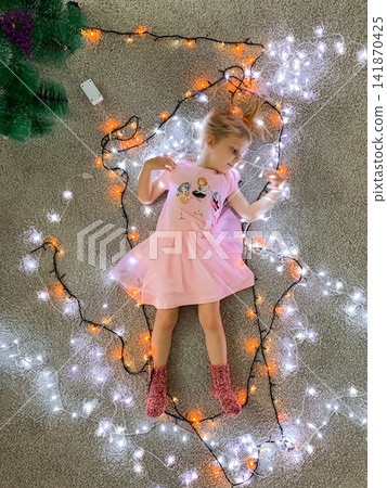 A little blonde girl in a pink dress lies on the floor surrounded by glowing orange and white Christmas fairy lights creating a magical holiday scene in a cozy home environment A little blonde girl in a pink dress lies on the floor surrounded by glowing orange and white Christmas fairy lights creating a magical holiday scene in a cozy home environment 141870425