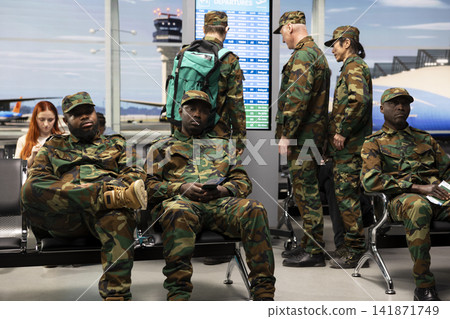 Military troops team in airport terminal sitting on bench, waiting for plane arrival. Multiracial army squad wearing camouflage uniforms bored in airport hub after flight got delayed Military troops team in airport terminal sitting on bench, waiting for plane arrival. Multiracial army squad wearing camouflage uniforms bored in airport hub after flight got delayed 141871749