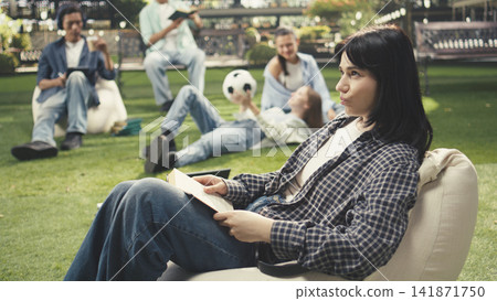Young woman student smiling while holding book on beanbag in sunny campus study area with classmates and greenery behind. Peer learning and reading study materials. Young woman student smiling while holding book on beanbag in sunny campus study area with classmates and greenery behind. Peer learning and reading study materials. 141871750