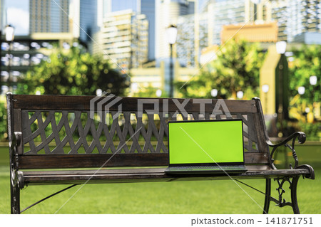 Chroma key replaceable screen on laptop in urban park with skyscrapers in background, empty wooden bench with generous copy space in sunlit peaceful landscape. Chroma key replaceable screen on laptop in urban park with skyscrapers in background, empty wooden bench with generous copy space in sunlit peaceful landscape. 141871751